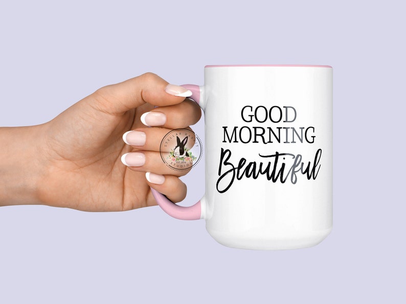Peut inclure: Une tasse en c&eacute;ramique blanche avec un bord et une poign&eacute;e roses. La tasse porte l'inscription "Good Morning Beautiful" en noir.