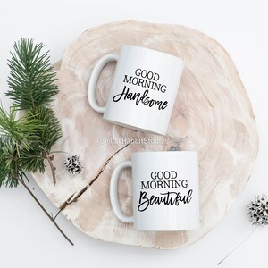 Peut inclure: Deux tasses en c&eacute;ramique blanche avec du texte noir. La tasse de gauche dit "Good Morning Handsome" et la tasse de droite dit "Good Morning Beautiful". Les tasses sont sur une surface en bois.