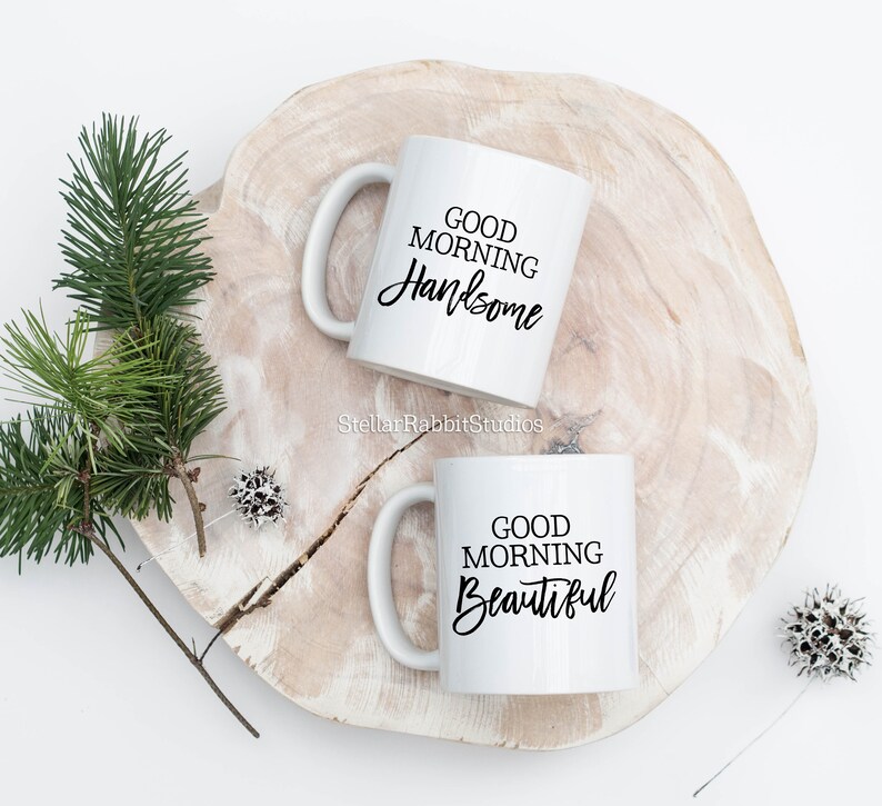 Peut inclure: Deux tasses en c&eacute;ramique blanche avec du texte noir. La tasse de gauche dit "Good Morning Handsome" et la tasse de droite dit "Good Morning Beautiful". Les tasses sont sur une surface en bois.