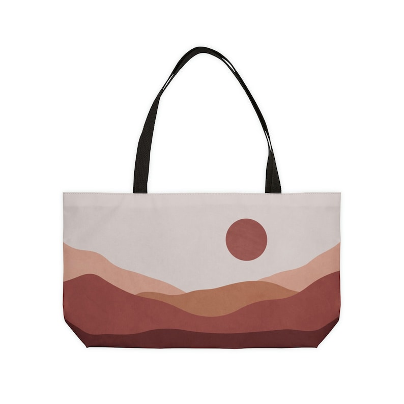 May include: A tote bag featuring a minimalist landscape design. The bag displays a warm color palette with a red sun and layered hills in shades of brown and peach. It has black handles.