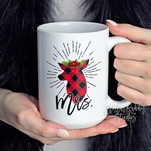 Peut inclure: Une tasse en c&eacute;ramique blanche avec une silhouette de cerf en plaid rouge et noir portant une couronne florale rouge et verte. Le cerf est entour&eacute; de rayons de soleil noirs et le mot "Mme" est imprim&eacute; sous le cerf.