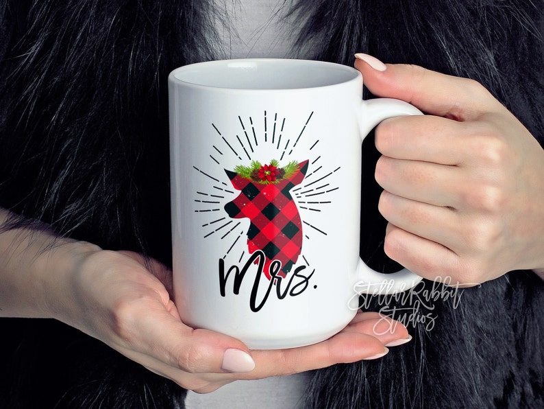 Peut inclure: Une tasse en c&eacute;ramique blanche avec une silhouette de cerf en plaid rouge et noir portant une couronne florale rouge et verte. Le cerf est entour&eacute; de rayons de soleil noirs et le mot "Mme" est imprim&eacute; sous le cerf.