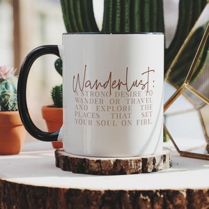 May include: White ceramic mug with a black handle. The mug has the text "Wanderlust: A strong desire to wander or travel and explore the places that set your soul on fire." printed in brown on the front.