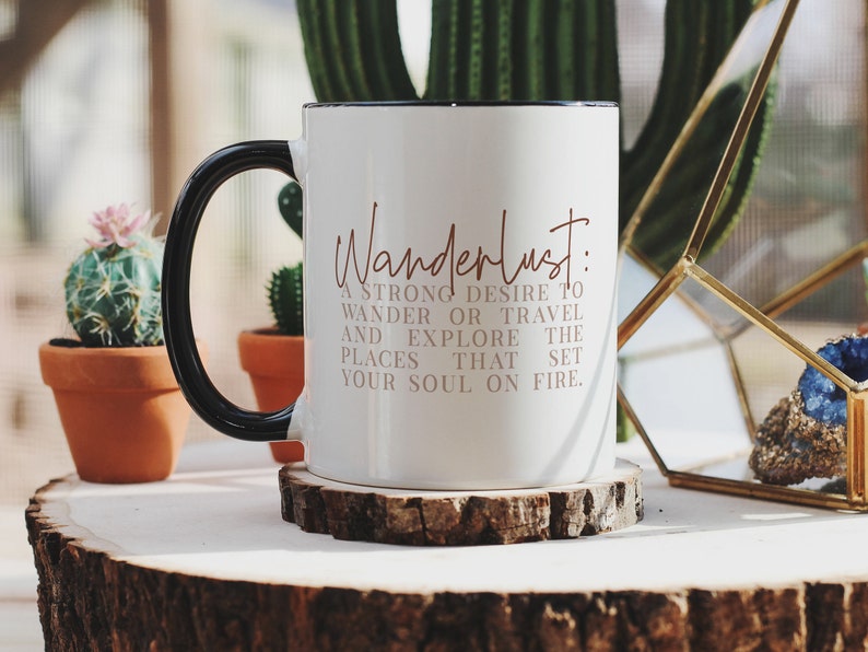 May include: White ceramic mug with a black handle. The mug has the text "Wanderlust: A strong desire to wander or travel and explore the places that set your soul on fire." printed in brown on the front.