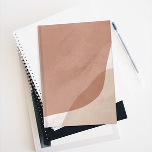 Pu&ograve; includere: Un quaderno marrone e beige con forme astratte e un design simile all'acquerello. Il quaderno &egrave; chiuso e si trova sopra un quaderno bianco a spirale.