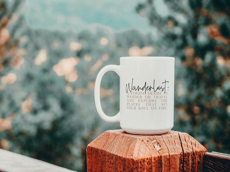 May include: White ceramic mug with a black inscription that reads "Wanderlust: A strong desire to wander or travel and explore the places that set your soul on fire". The mug is sitting on a wooden surface with a blurred background of green foliage.