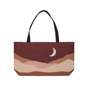 May include: A tote bag featuring a landscape design with a crescent moon. The bag has a dark red background with layered tan and brown hills. It has black handles and is likely made of fabric.