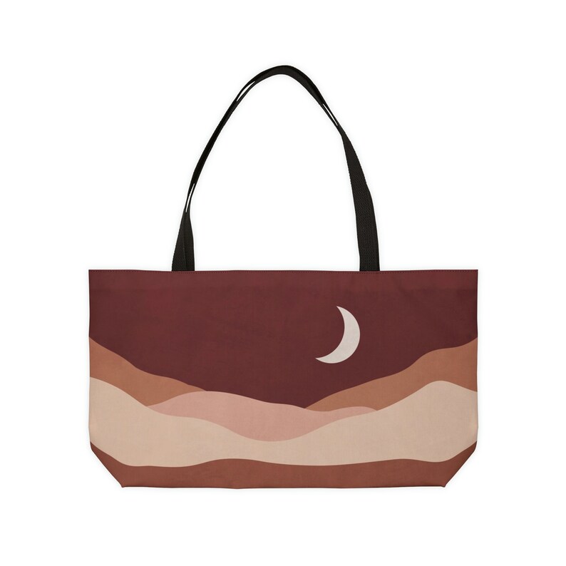 May include: A tote bag featuring a landscape design with a crescent moon. The bag has a dark red background with layered tan and brown hills. It has black handles and is likely made of fabric.