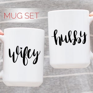 Peut inclure: Deux tasses en c&eacute;ramique blanche avec une calligraphie noire. La tasse de gauche dit "wifey" avec un petit c&oelig;ur au-dessus du "y". La tasse de droite dit "hubby".