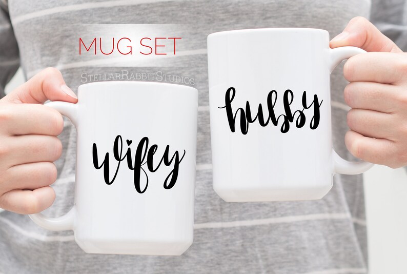 Peut inclure: Deux tasses en c&eacute;ramique blanche avec une calligraphie noire. La tasse de gauche dit "wifey" avec un petit c&oelig;ur au-dessus du "y". La tasse de droite dit "hubby".