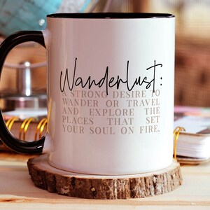 May include: White ceramic mug with a black handle and black rim. The mug has the text "Wanderlust: A strong desire to wander or travel and explore the places that set your soul on fire." printed in black.