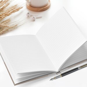 Pu&ograve; includere: Un quaderno a copertina rigida aperto con una penna d'argento su una superficie bianca. Il quaderno &egrave; aperto su una pagina bianca e la penna &egrave; appoggiata sul quaderno.