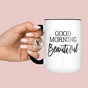 Peut inclure: Une tasse en c&eacute;ramique blanche avec un bord et une poign&eacute;e noirs. La tasse porte l'inscription noire "Good Morning Beautiful".