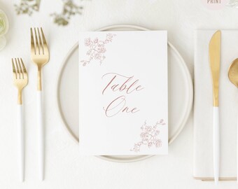 May include: A white table number card with the text "Table One" in a delicate script font, adorned with floral corner accents. Gold and white flatware is arranged around the card, suggesting a wedding or event setting. The words "READY TO PRINT" are in the upper right corner.