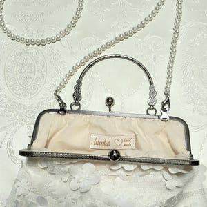 May include: A white clutch purse with a pearl chain strap. The purse is made of white lace with 3D flower embellishments. The purse has a silver clasp and a silver handle. The inside of the purse is lined with a cream-colored fabric. The purse has a label that says "Lebenslust hand made".