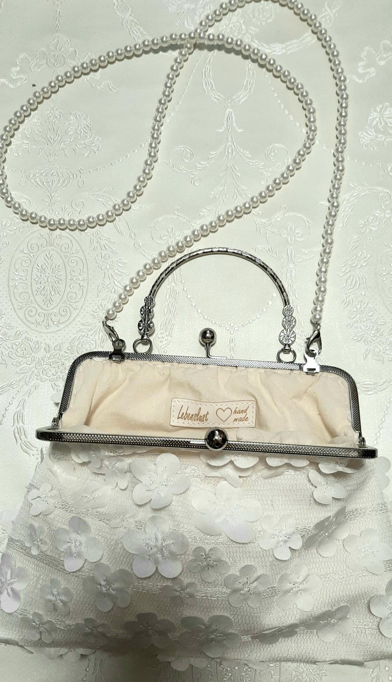 May include: A white clutch purse with a pearl chain strap. The purse is made of white lace with 3D flower embellishments. The purse has a silver clasp and a silver handle. The inside of the purse is lined with a cream-colored fabric. The purse has a label that says "Lebenslust hand made".