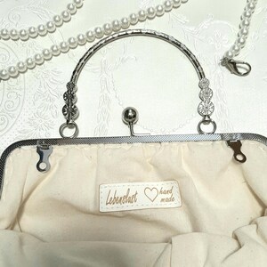 May include: A white clutch purse with a silver frame and a silver handle. The purse is decorated with white flowers and has a label that reads "Lebenslust hand made".