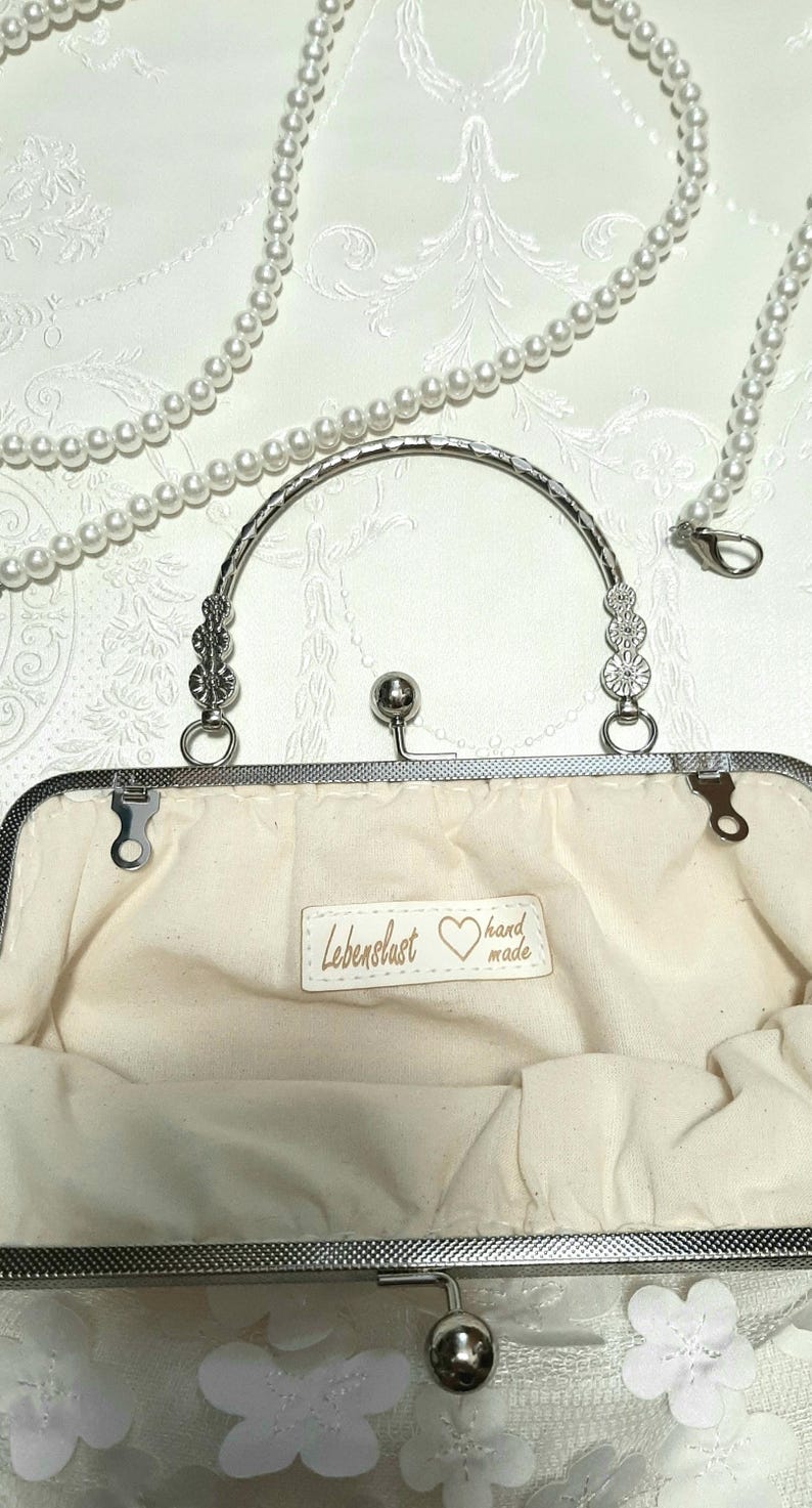 May include: A white clutch purse with a silver frame and a silver handle. The purse is decorated with white flowers and has a label that reads "Lebenslust hand made".