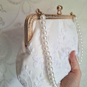 Cremefarbene Clutch mit goldfarbenem Verschluss und Perlenkette. Der Stoff ist mit einem zarten Blumenmuster bestickt. Die Clutch hat eine weiche, abgerundete Form.