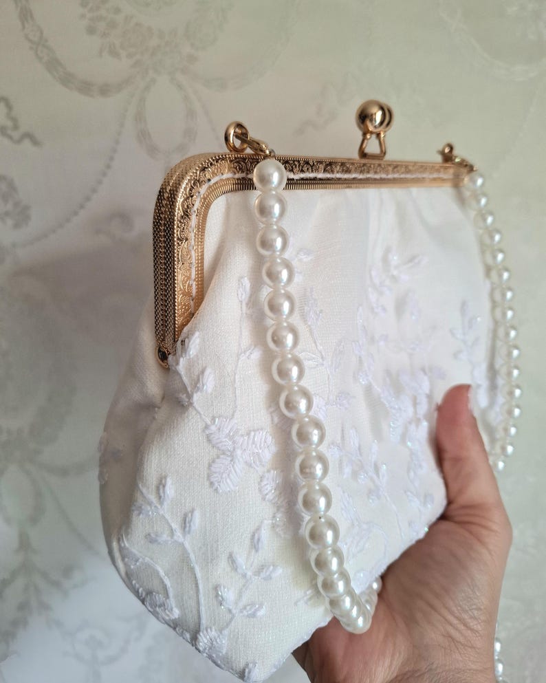 Cremefarbene Clutch mit goldfarbenem Verschluss und Perlenkette. Der Stoff ist mit einem zarten Blumenmuster bestickt. Die Clutch hat eine weiche, abgerundete Form.