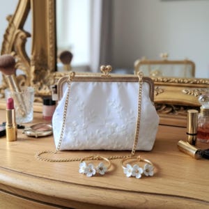 Eine weiße, floral bestickte Braut Clutch mit goldener Kette und Verschluss. Zwei goldene Creolen mit weißen Blumenakzenten liegen vor der Clutch. Make-up-Artikel befinden sich im Hintergrund.