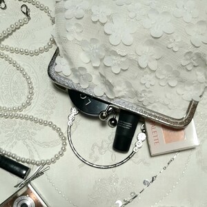 May include: A white clutch purse with a floral design. The purse has a silver clasp and a silver chain handle. The purse is open and there are makeup items inside, including a black tube of lipstick, a black compact, and a pink compact with the word "Palette" on it.