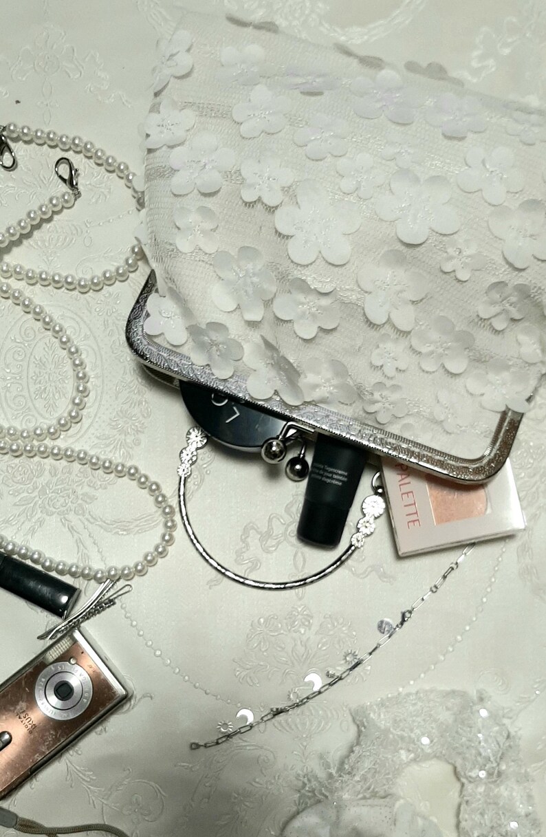 May include: A white clutch purse with a floral design. The purse has a silver clasp and a silver chain handle. The purse is open and there are makeup items inside, including a black tube of lipstick, a black compact, and a pink compact with the word "Palette" on it.