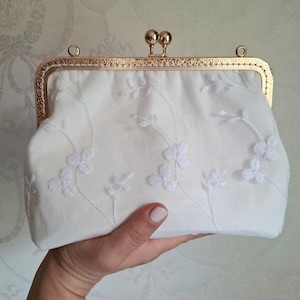 May include: A white clutch with a gold-colored frame and clasp. The clutch is embroidered with a delicate floral pattern. The frame has an ornate design. The clutch is held in a person's hand.