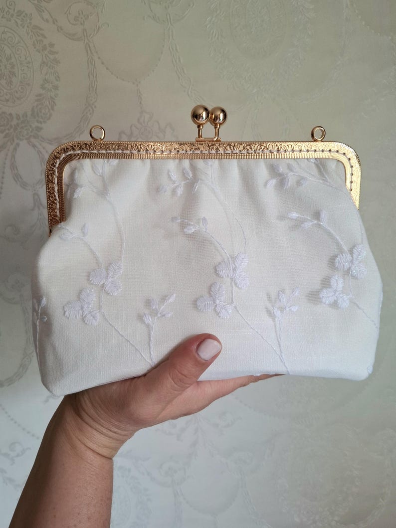 May include: A white clutch with a gold-colored frame and clasp. The clutch is embroidered with a delicate floral pattern. The frame has an ornate design. The clutch is held in a person's hand.