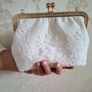 Eine cremefarbene Brauttasche mit einem goldfarbenen Rahmen und Verschluss. Die Tasche ist mit weißen Blumenstickereien verziert. Die Clutch wird in einer Hand gehalten und unterstreicht ihr elegantes Design.