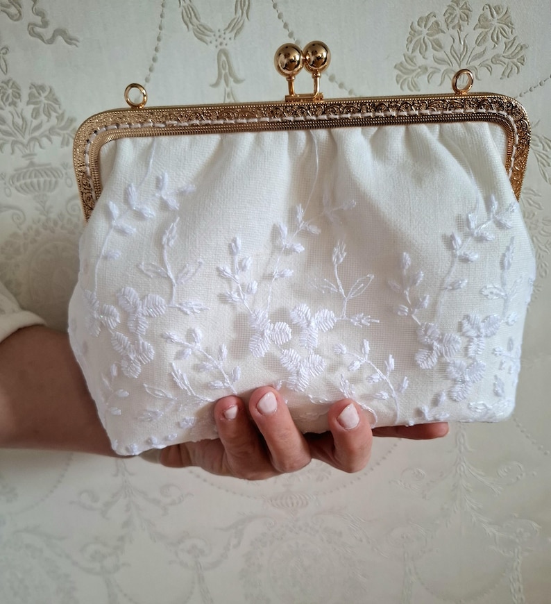 Eine cremefarbene Brauttasche mit einem goldfarbenen Rahmen und Verschluss. Die Tasche ist mit weißen Blumenstickereien verziert. Die Clutch wird in einer Hand gehalten und unterstreicht ihr elegantes Design.