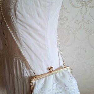 Eine cremefarbene Clutch mit floraler Stickerei, einem goldfarbenen Verschluss und einem perlenbesetzten Riemen. Die Tasche wird vor einem weißen Kleid gehalten, was auf einen formellen Anlass hindeutet. Das Design ist zart und feminin.