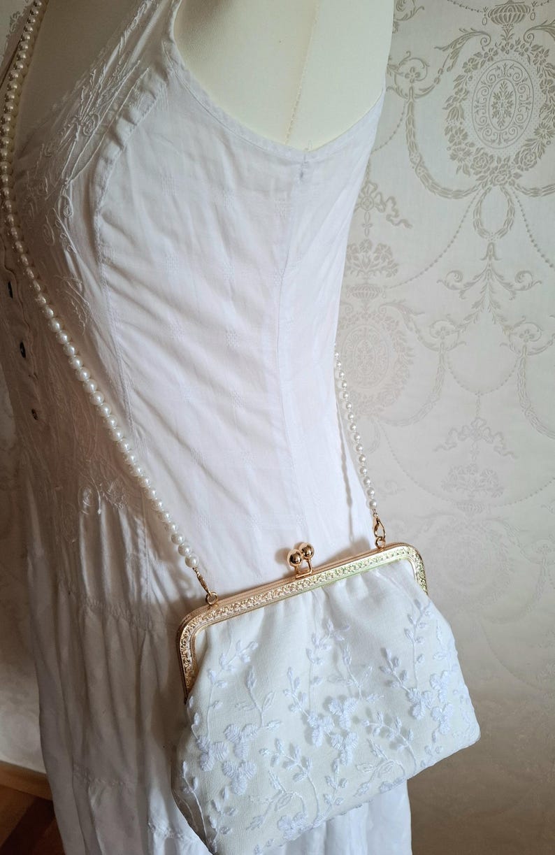 Eine cremefarbene Clutch mit floraler Stickerei, einem goldfarbenen Verschluss und einem perlenbesetzten Riemen. Die Tasche wird vor einem weißen Kleid gehalten, was auf einen formellen Anlass hindeutet. Das Design ist zart und feminin.