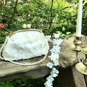 May include: A white floral clutch purse with a pearl chain strap. The purse is sitting on a wooden bench with a white floral garland and a candle holder.