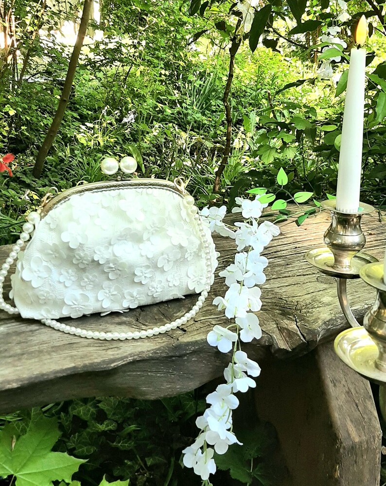 May include: A white floral clutch purse with a pearl chain strap. The purse is sitting on a wooden bench with a white floral garland and a candle holder.