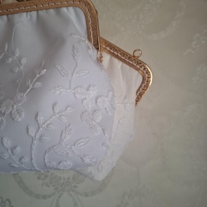 May include: Two white, vintage-style purses with gold-toned frames. The purses feature delicate floral embroidery and a clasp closure. The background is a light beige color with a subtle pattern.