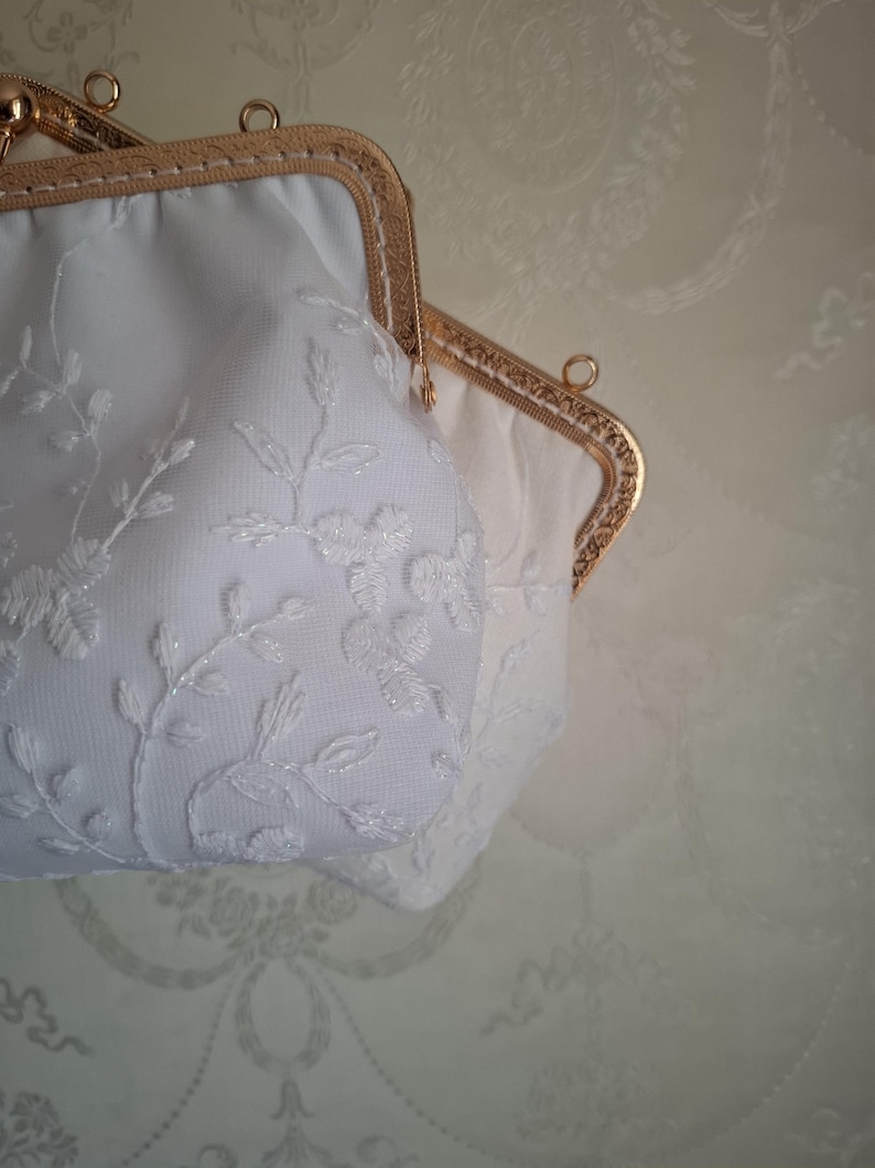 May include: Two white, vintage-style purses with gold-toned frames. The purses feature delicate floral embroidery and a clasp closure. The background is a light beige color with a subtle pattern.
