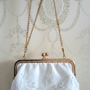 Weiße Satin-Clutch mit goldfarbenem Rahmen und Kette. Die Clutch hat zarte weiße Blumenstickereien. Der Verschluss ist ein goldfarbener Kugelverschluss. Ein klassisches Accessoire für Hochzeit und  besondere Anlässe.