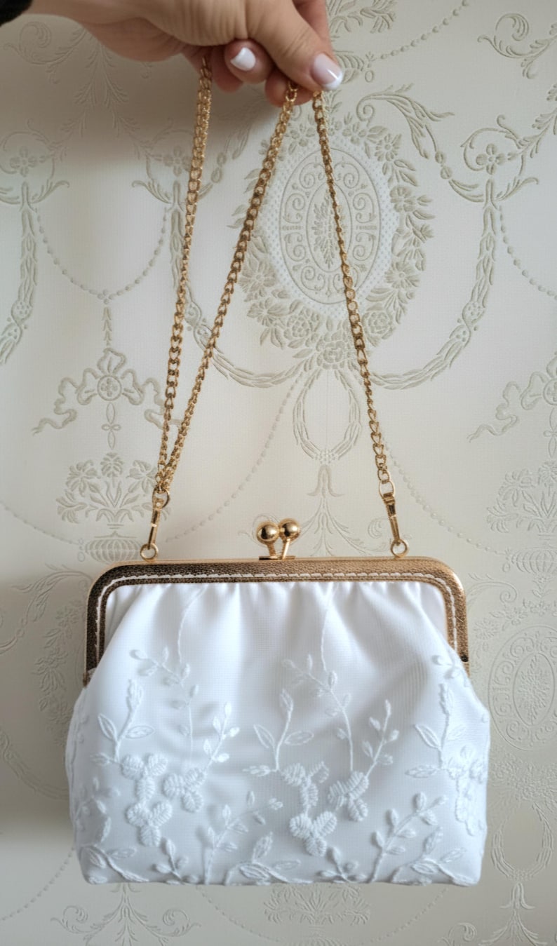 Weiße Satin-Clutch mit goldfarbenem Rahmen und Kette. Die Clutch hat zarte weiße Blumenstickereien. Der Verschluss ist ein goldfarbener Kugelverschluss. Ein klassisches Accessoire für Hochzeit und  besondere Anlässe.