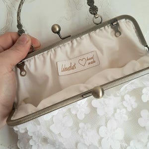 May include: White floral clutch purse with a silver metal frame and handle. The purse has a label that says "Lebenslust hand made".