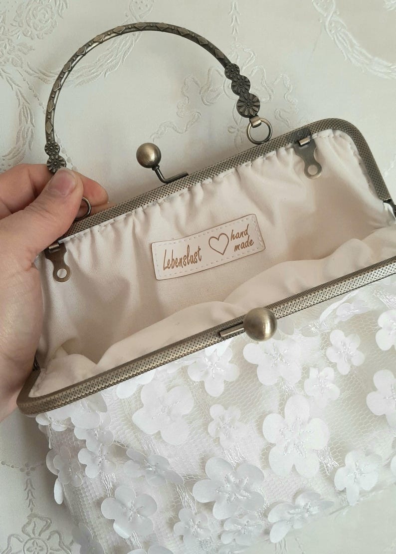 May include: White floral clutch purse with a silver metal frame and handle. The purse has a label that says "Lebenslust hand made".