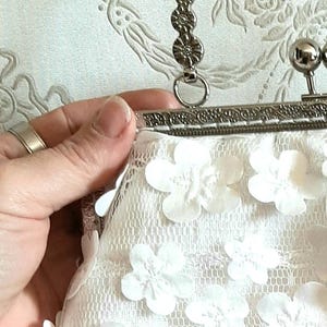 May include: A white clutch purse with a silver clasp and handle. The purse is made of lace and has white flower embellishments.