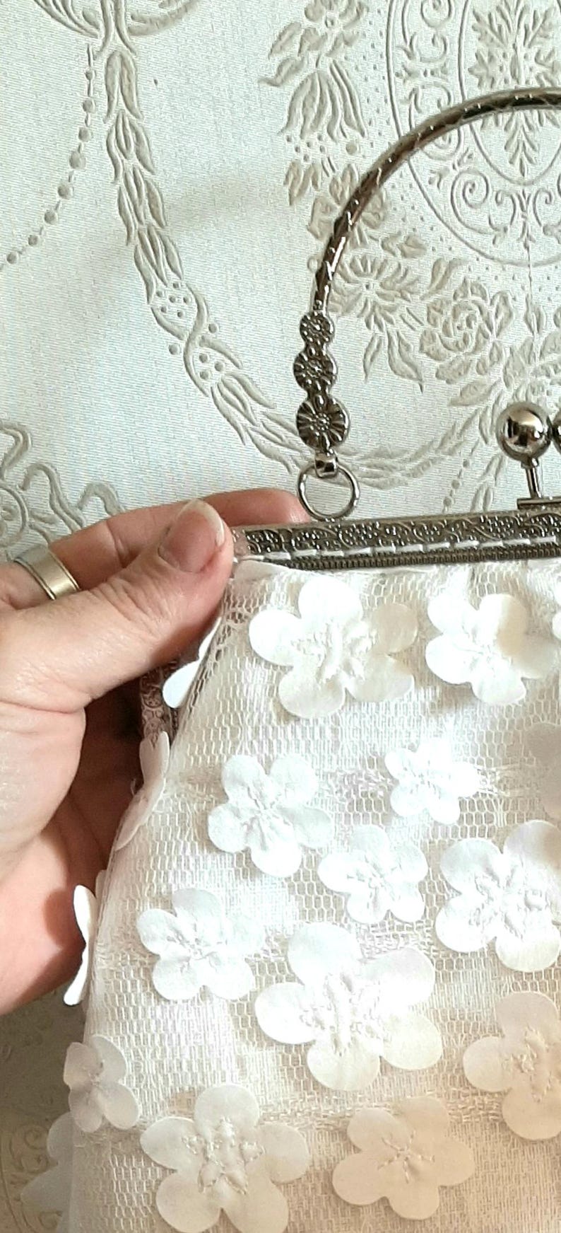 May include: A white clutch purse with a silver clasp and handle. The purse is made of lace and has white flower embellishments.