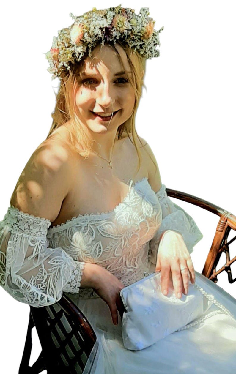 May include: A woman wearing a white off-the-shoulder dress with floral lace detailing and long, sheer sleeves. She is wearing a flower crown and holding a white clutch. The dress appears to be a bridal or formal gown.