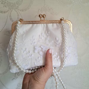 ivory farbene Clutch mit goldfarbenem Verschluss und Perlenriemen. Die Clutch hat zarte weiße Blumenstickereien. Der Perlenriemen ist über die Clutch drapiert. Der Verschluss hat eine abgerundete rechteckige Form.