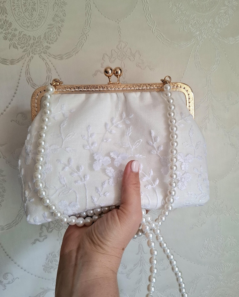 ivory farbene Clutch mit goldfarbenem Verschluss und Perlenriemen. Die Clutch hat zarte weiße Blumenstickereien. Der Perlenriemen ist über die Clutch drapiert. Der Verschluss hat eine abgerundete rechteckige Form.