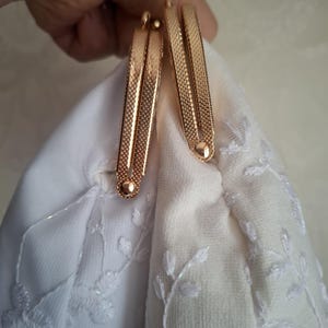 May include: Close-up of a white fabric purse with a floral embroidered design. The purse features a gold-colored metal clasp with a textured pattern. The clasp is held open, revealing the purse's interior.