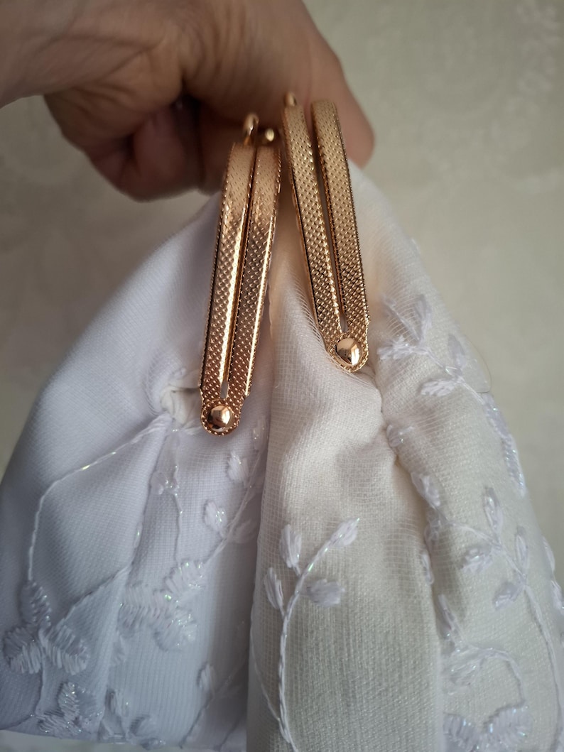 May include: Close-up of a white fabric purse with a floral embroidered design. The purse features a gold-colored metal clasp with a textured pattern. The clasp is held open, revealing the purse's interior.