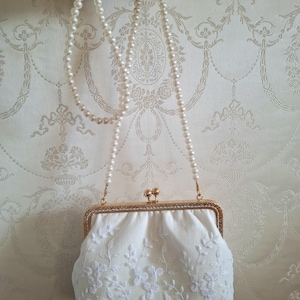 Eine weiße, floral Bestickte Abendtasche mit goldfarbenem Rahmen und einer perlenbesetzten Kette. Die Clutch hat einen Kiss-Lock-Verschluss und befindet sich vor einem gemusterten Hintergrund.