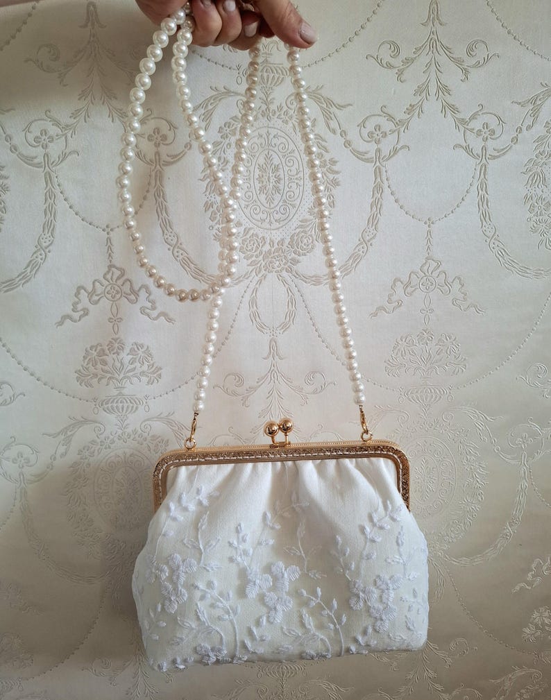Eine weiße, floral Bestickte Abendtasche mit goldfarbenem Rahmen und einer perlenbesetzten Kette. Die Clutch hat einen Kiss-Lock-Verschluss und befindet sich vor einem gemusterten Hintergrund.